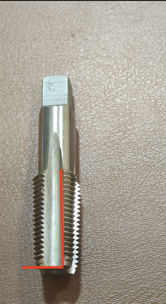 A hand tap with a square drive shank and straight flutes, with a red vertical line marking the flute angle running parallel to the tap body, showing threads and a pointed tip.